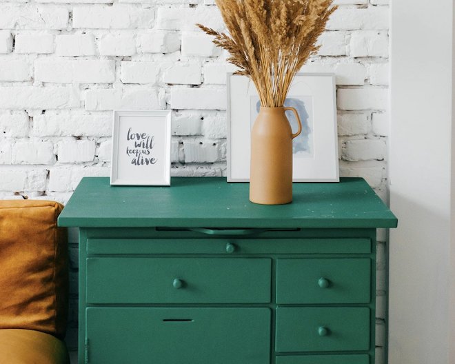 A green wooden dresser with a framed 'love will keep us alive' picture and a vase of wheat stalks, against a white brick wall.